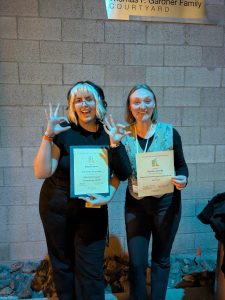 Kelsie Ramos and Keelin Connolly standing in front of a brick wall holding their certificates from the Kennedy Center American College Theatre Festival Conference.