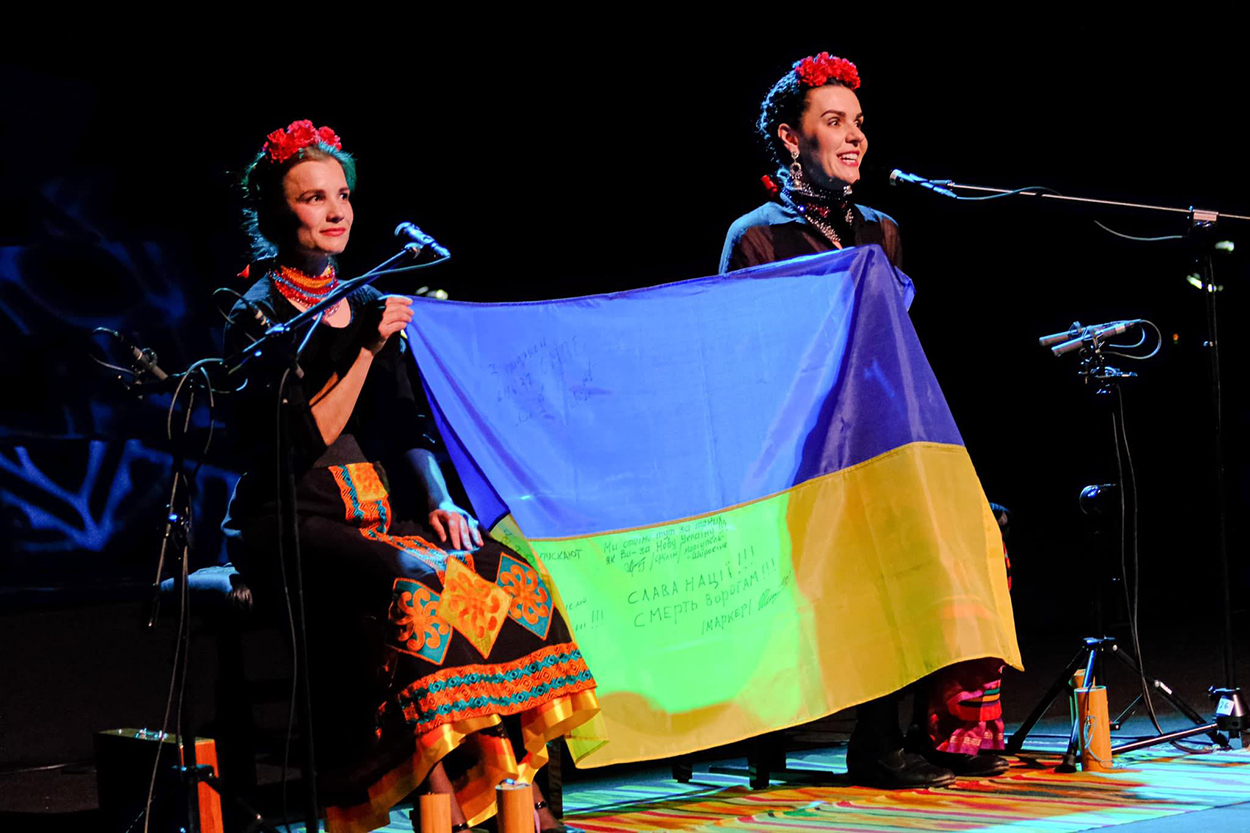 Kurbasy-photo-courtesy-of-the-artists-lr two women on stage with microphones hold up an Ukrainian flag